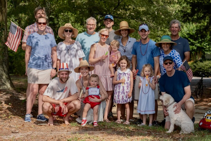 Multigenerational family group posing outdoors in a park for a patriotic gathering, with American flags and children in front.