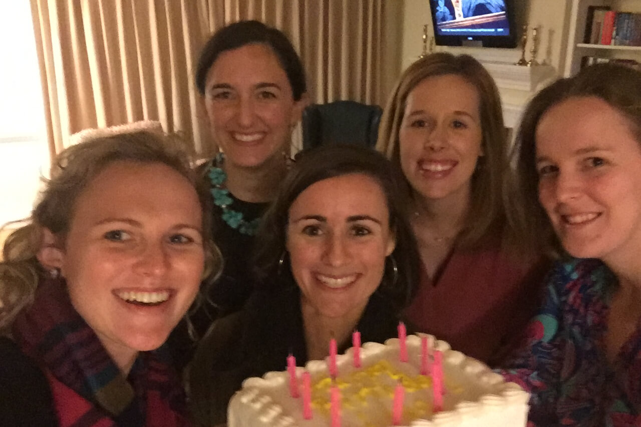 Five women smiling around a birthday cake with pink candles indoors.