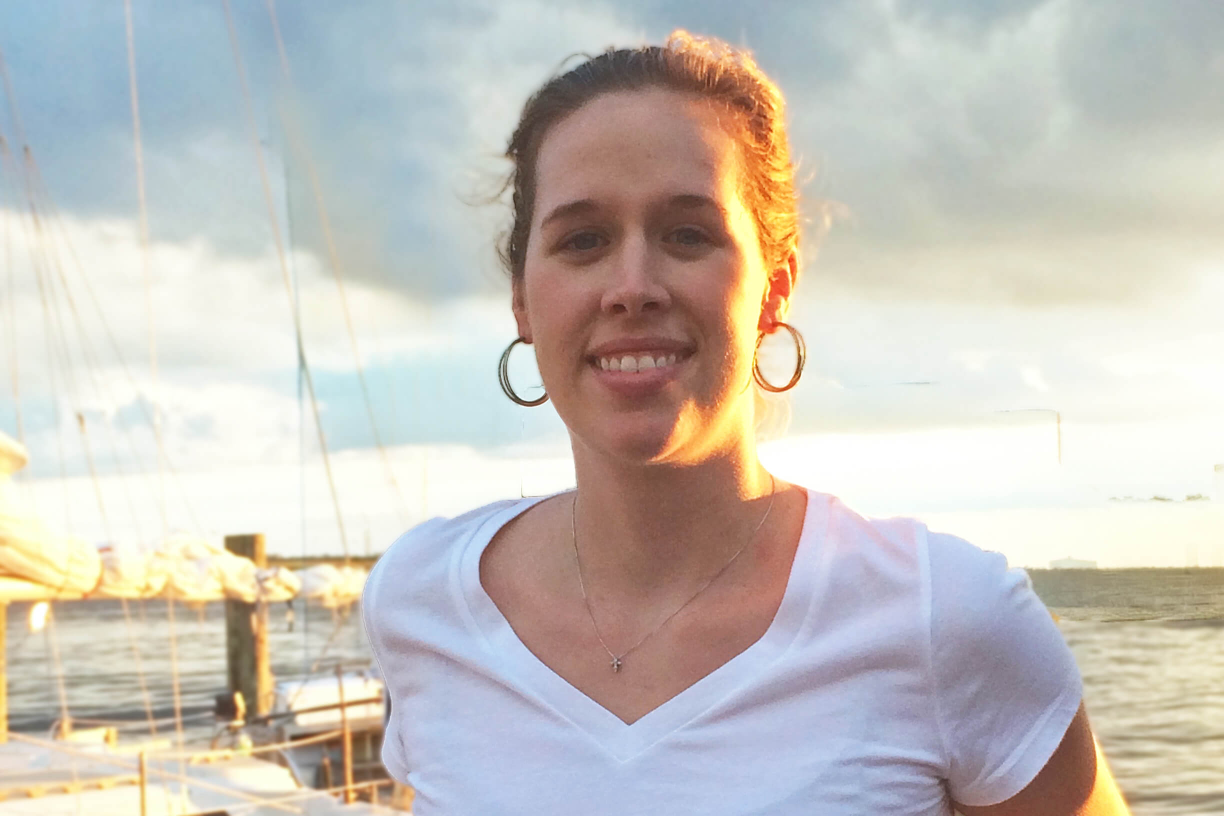 Smiling woman in a white shirt with hoop earrings, sunset-lit marina in the background and boats in the water.