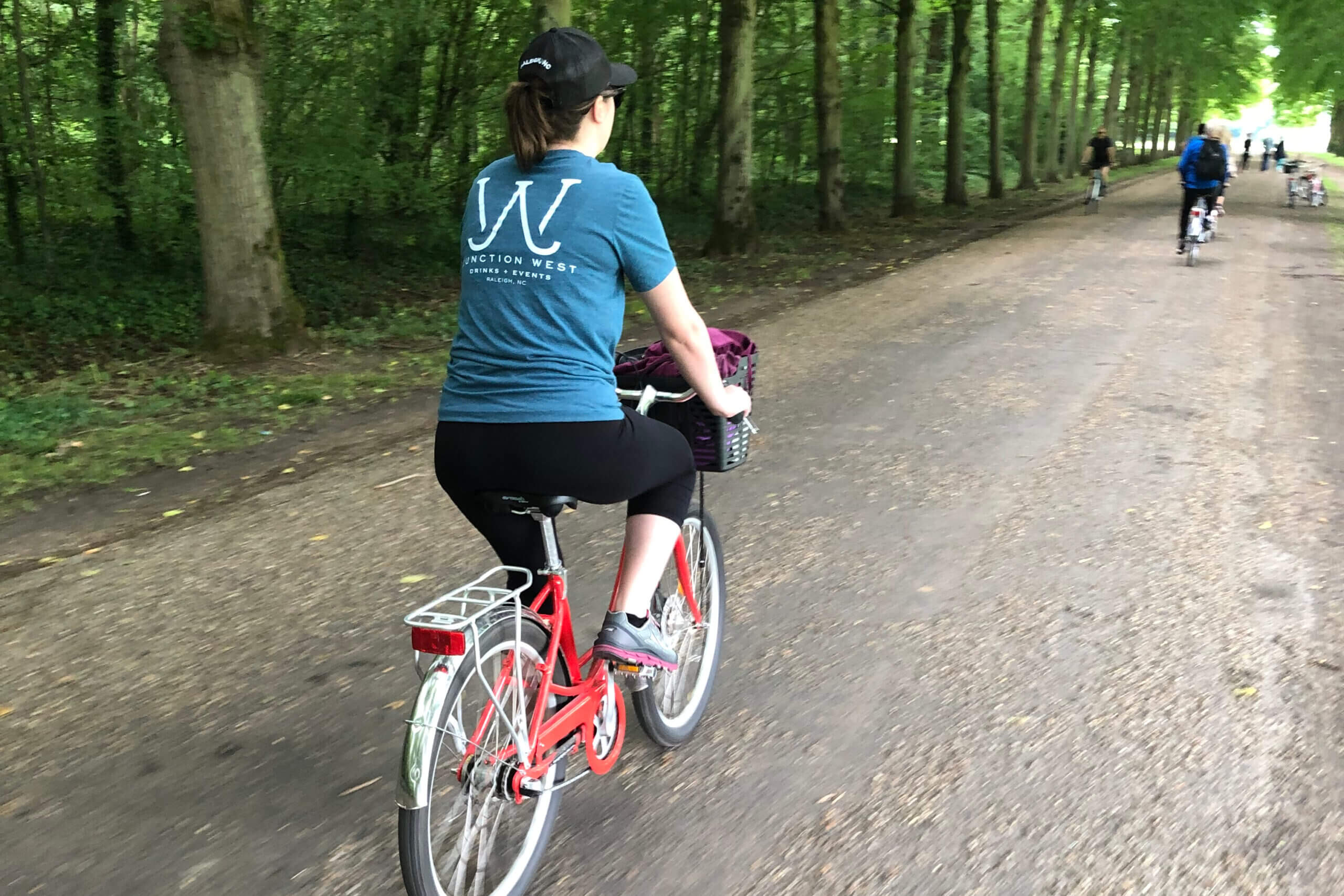 Cyclist wearing a blue shirt rides a red bike along a tree-lined dirt path; other riders are ahead in the distance.