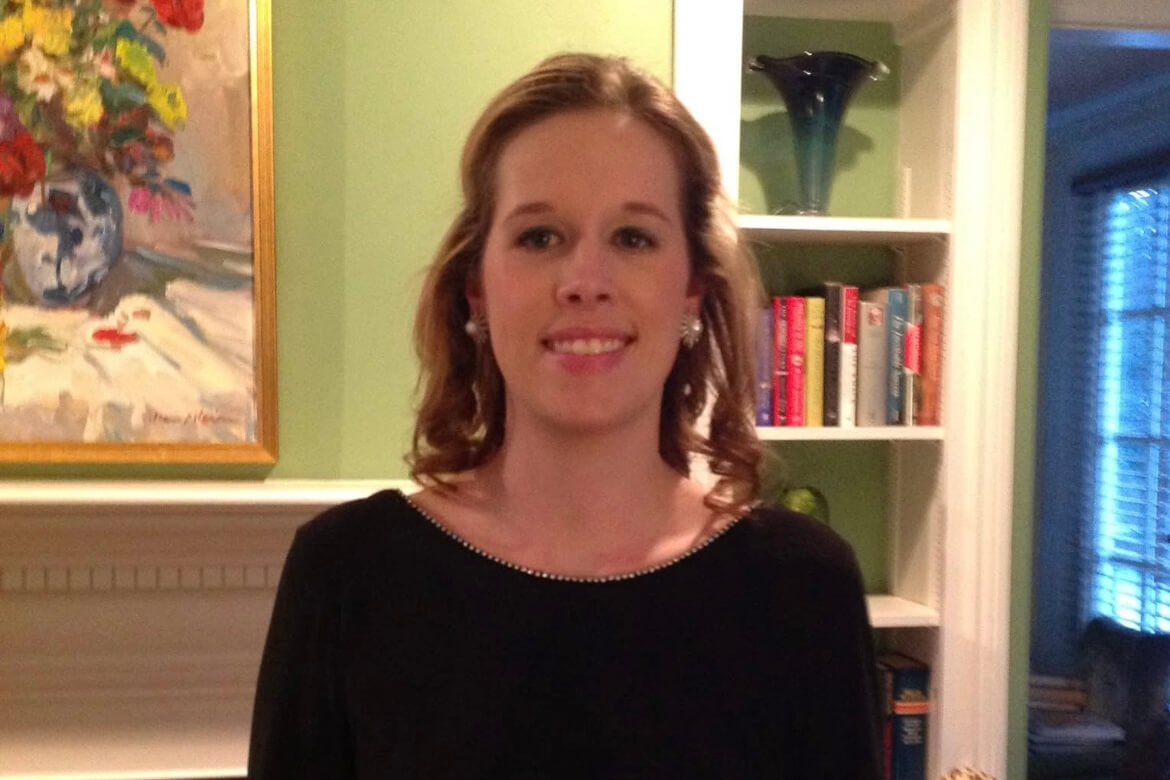 Woman smiling indoors with shoulder-length brown hair, wearing a black top, standing in front of a green wall and bookshelf.