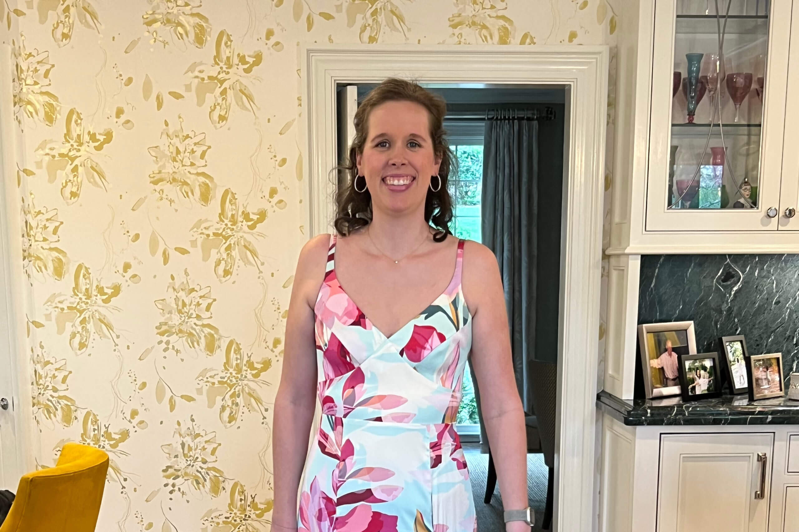 Smiling woman in a colorful floral dress standing indoors, with a display cabinet of glassware and photos visible behind her.