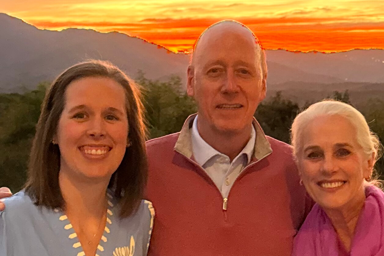Three adults smiling, posing together outdoors at sunset with a mountain range in the background.