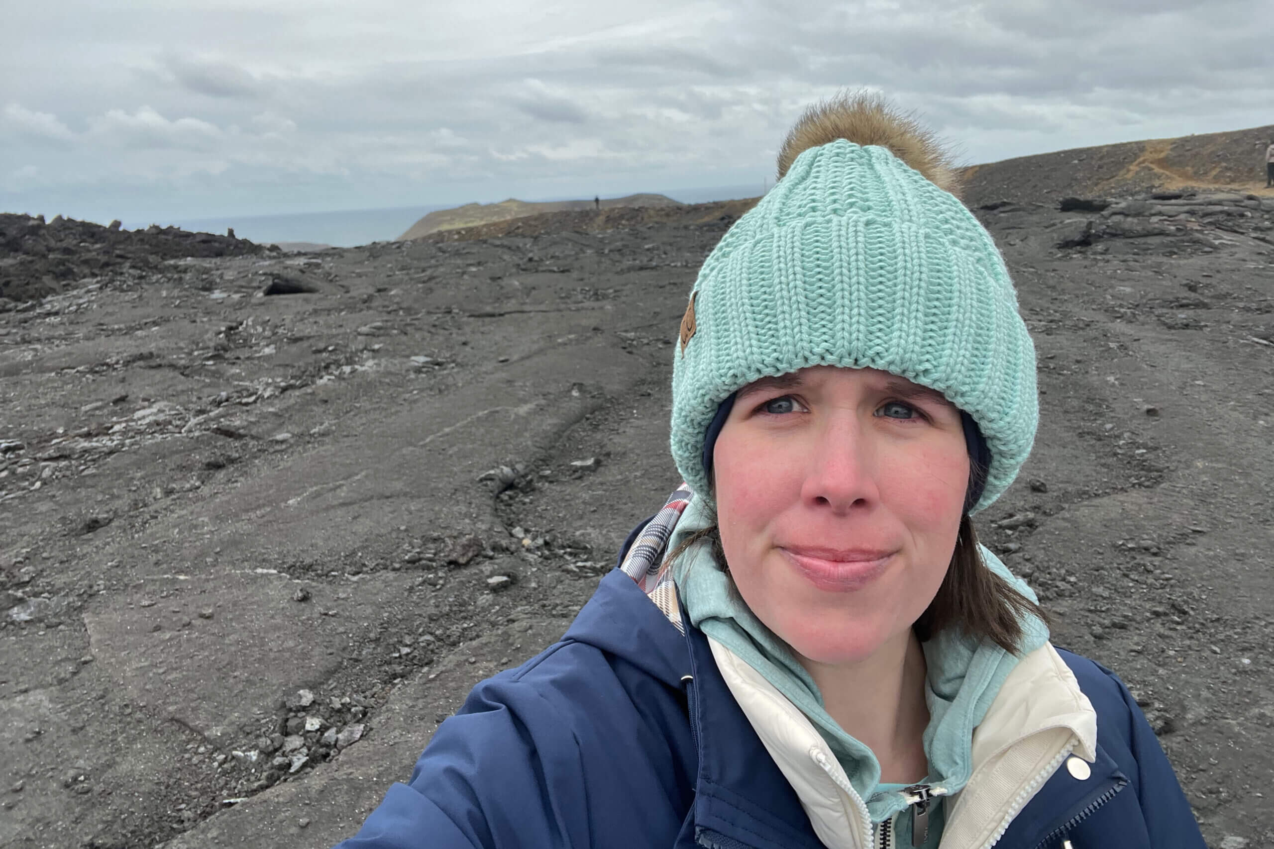 Selfie of a person wearing a mint green knit hat with a fur pom-pom, on a rocky volcanic field under a cloudy sky.