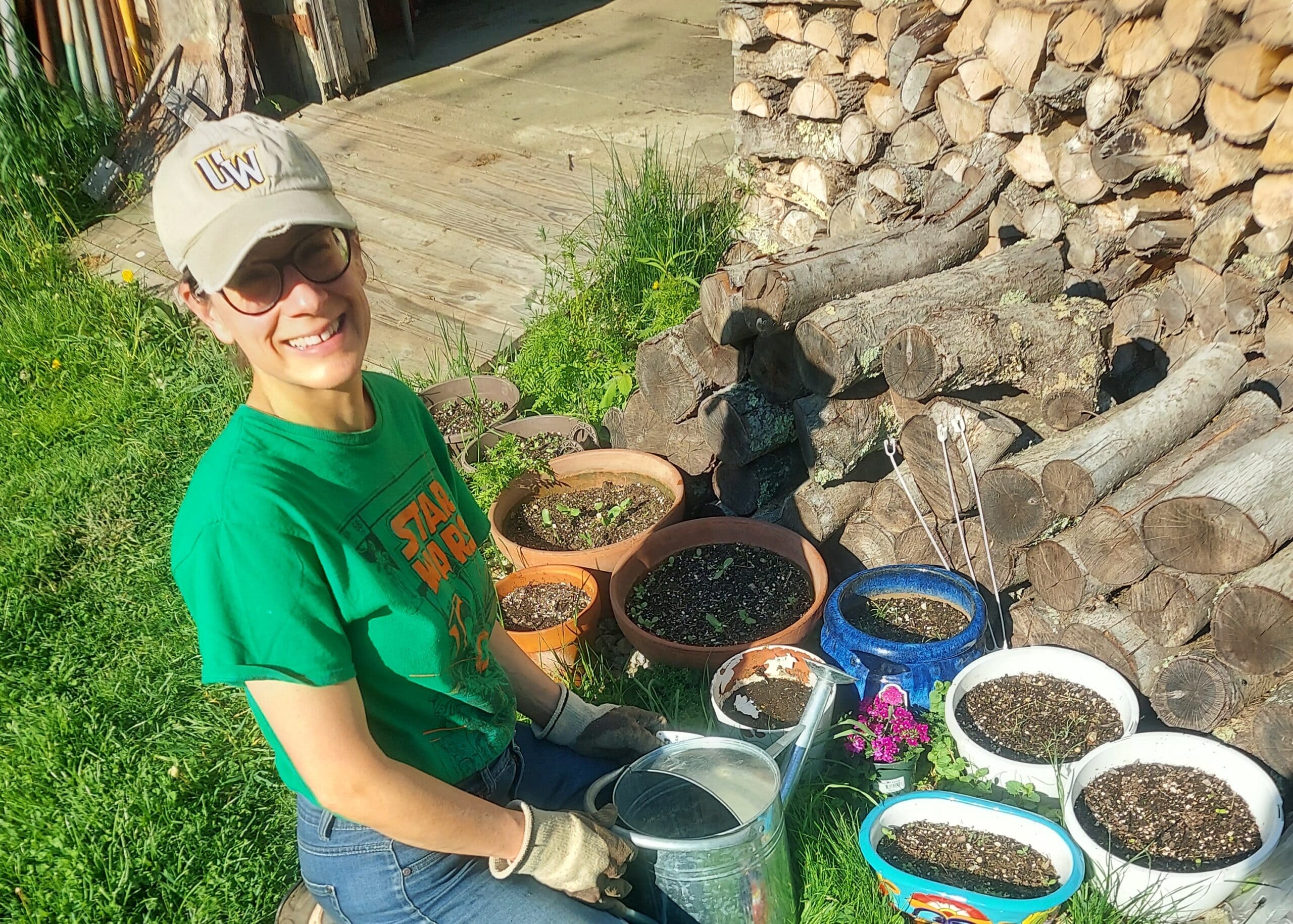 Person in a green shirt and cap smiles while gardening with several pots of soil and young plants; a stacked woodpile is in the background.