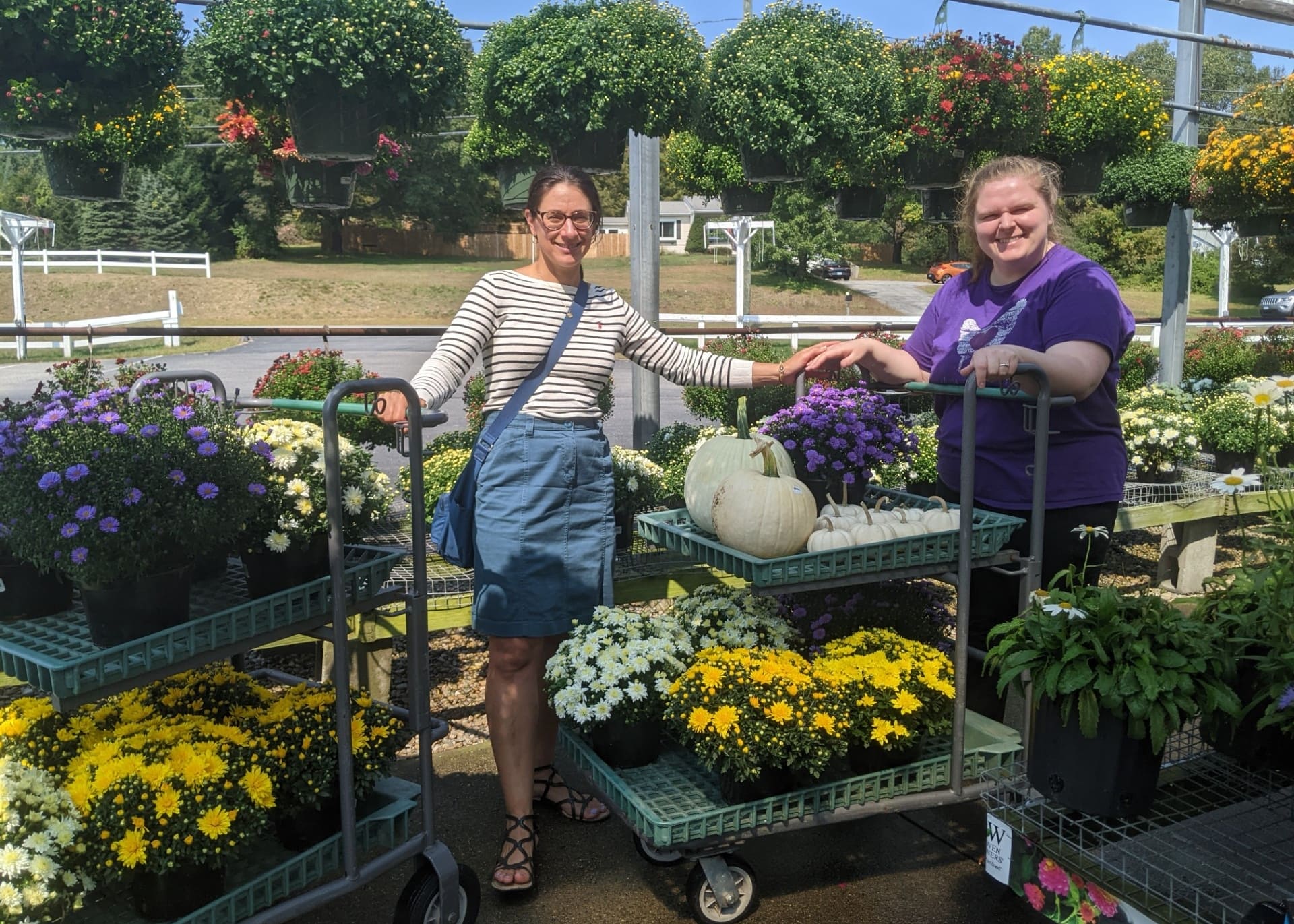 Two women at a sunny flower stand, smiling, with carts of colorful potted mums and a white pumpkin on display between them.
