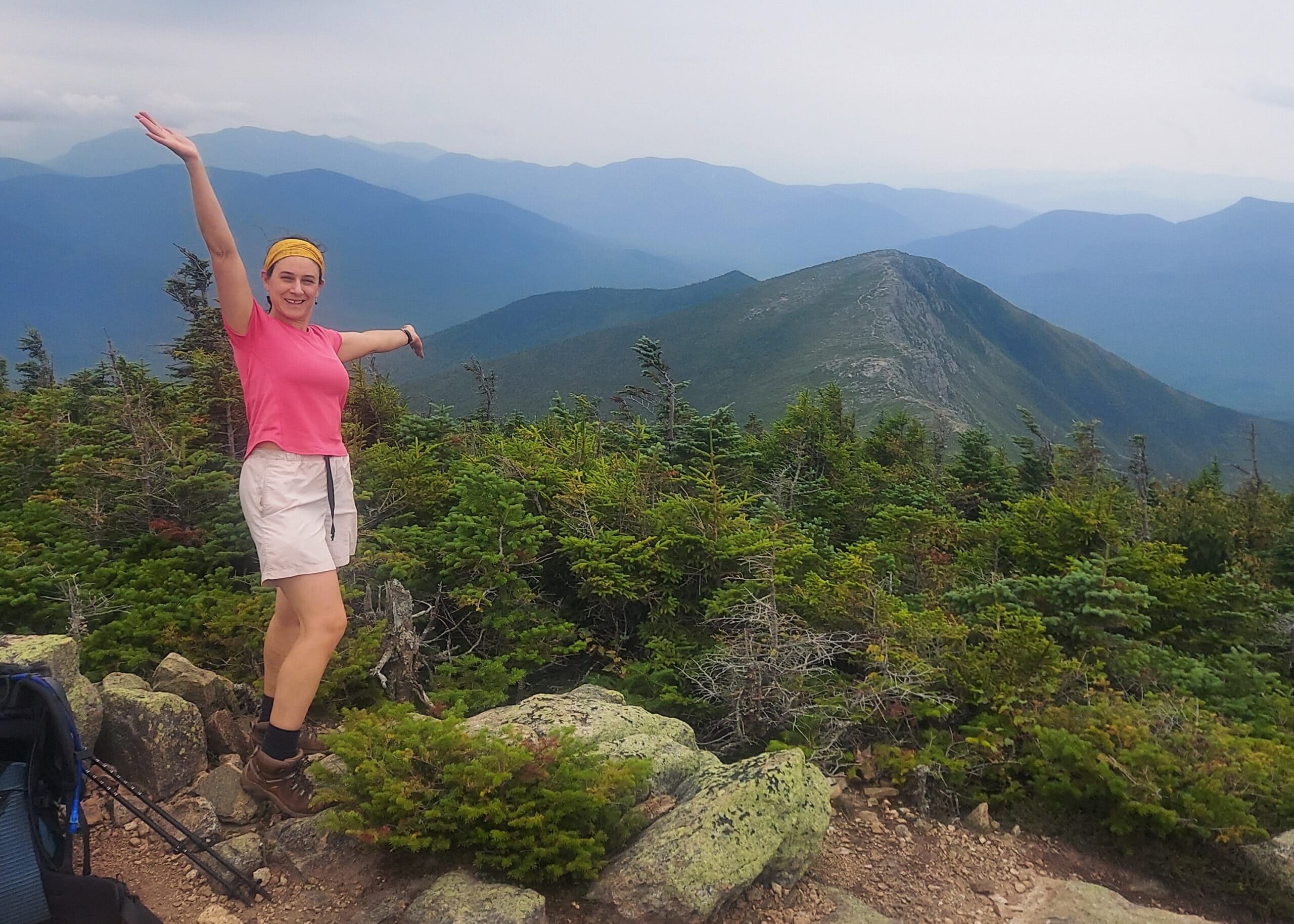 Woman hiker in a pink shirt and yellow headband raises both arms on a rocky mountain summit, with evergreen trees and blue ridges in the background, celebrating.