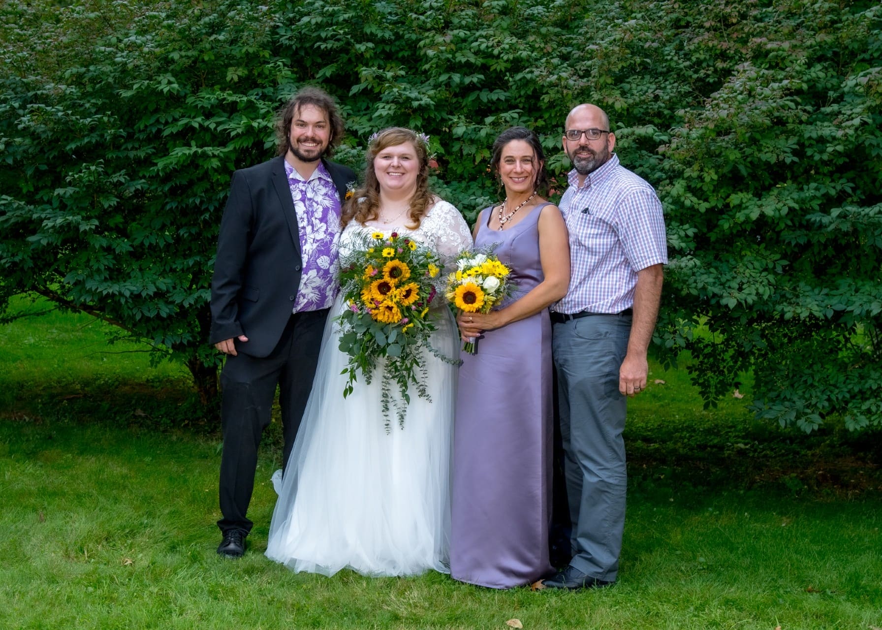 Group of four adults posing outdoors at a wedding, with the bride holding a bouquet and two guests in lavender dresses, green shrub背景 behind them.