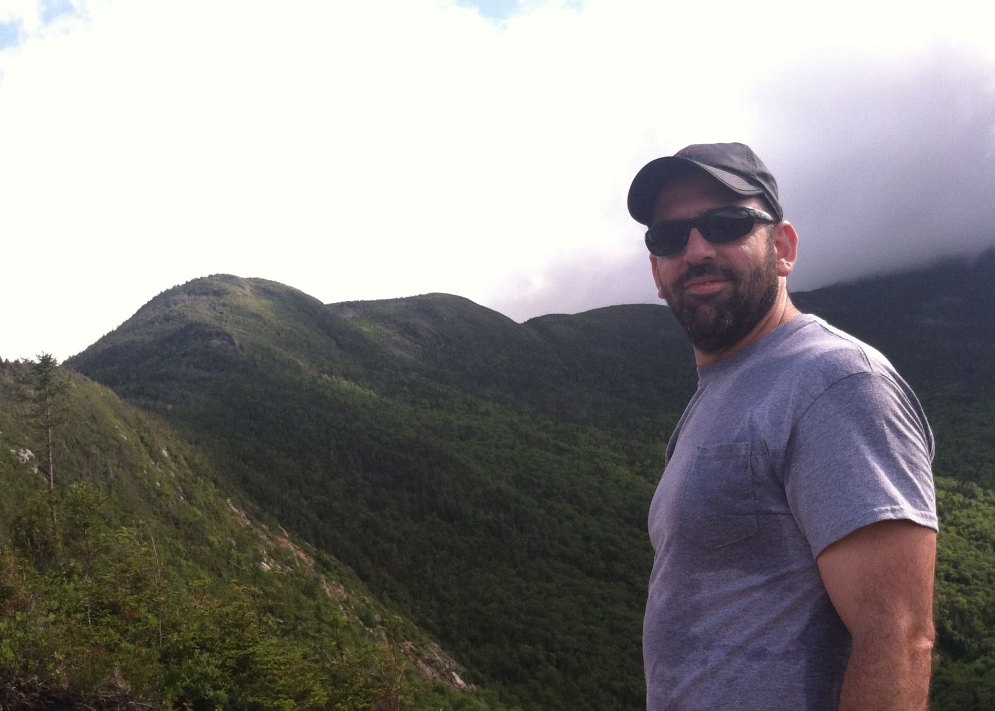 Man wearing a cap and sunglasses, standing on a green mountainside.
