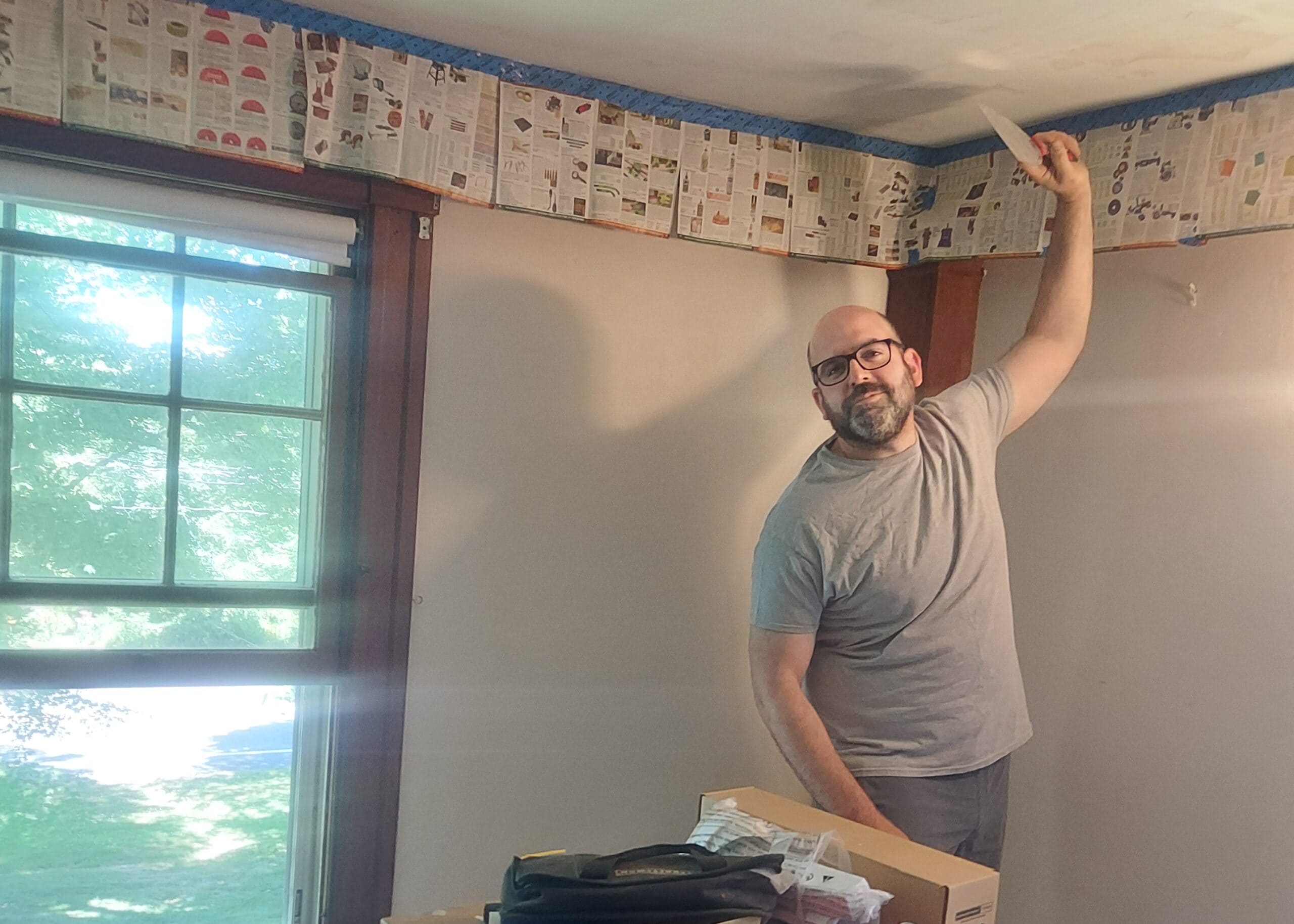 Man with glasses and a beard applies newspaper wallpaper to a ceiling, arm raised as he fits a page edge.