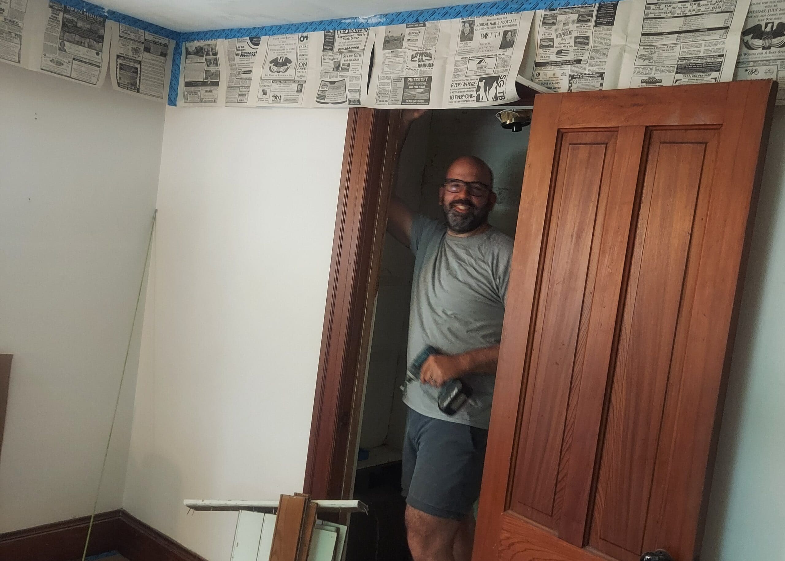 Man wearing glasses and a gray t-shirt stands in a doorway, holding a drill, in a room under renovation.