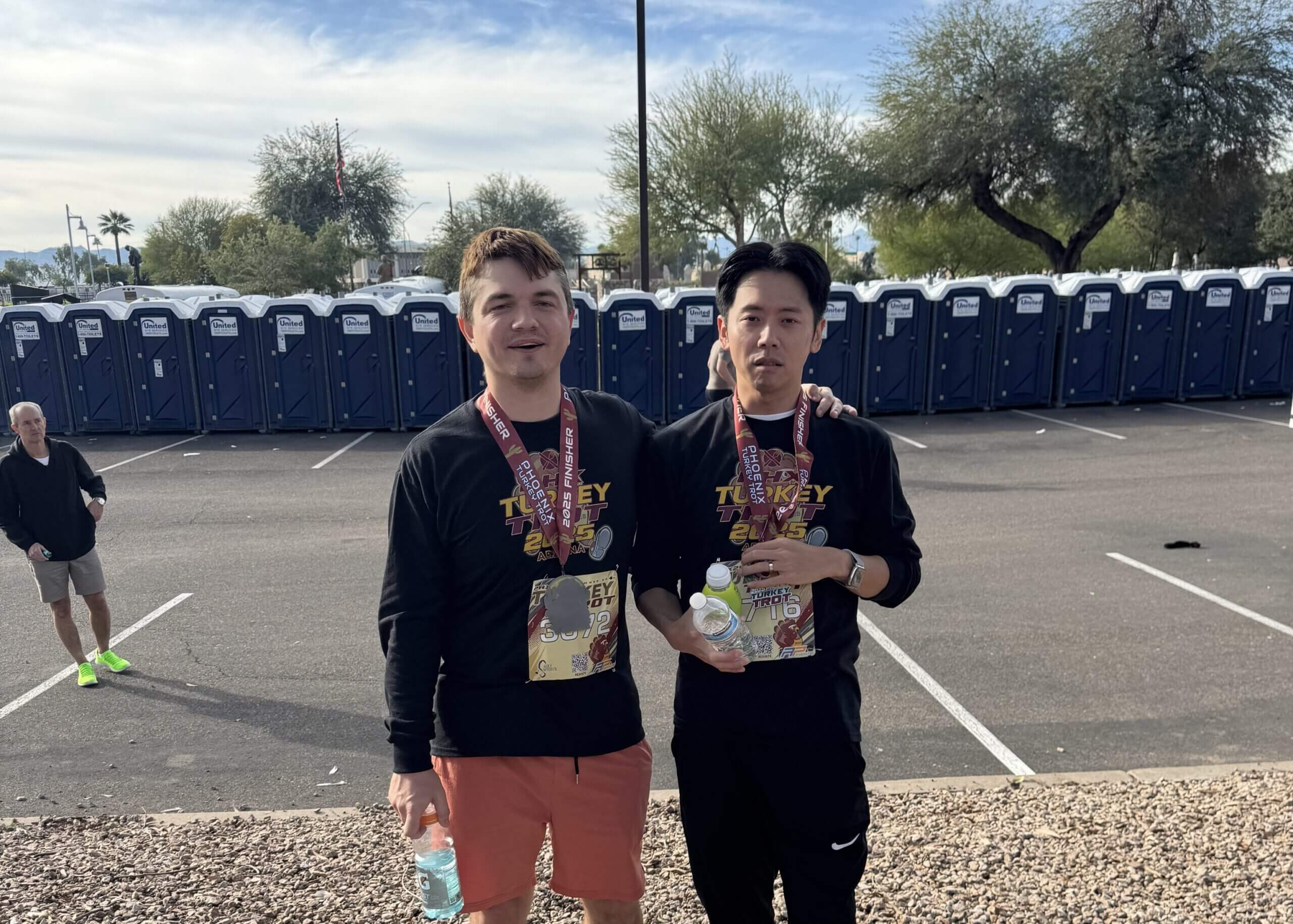 Two male runners posing for a photo after a race, medals around their necks, with portable toilets lining a parking lot behind them.