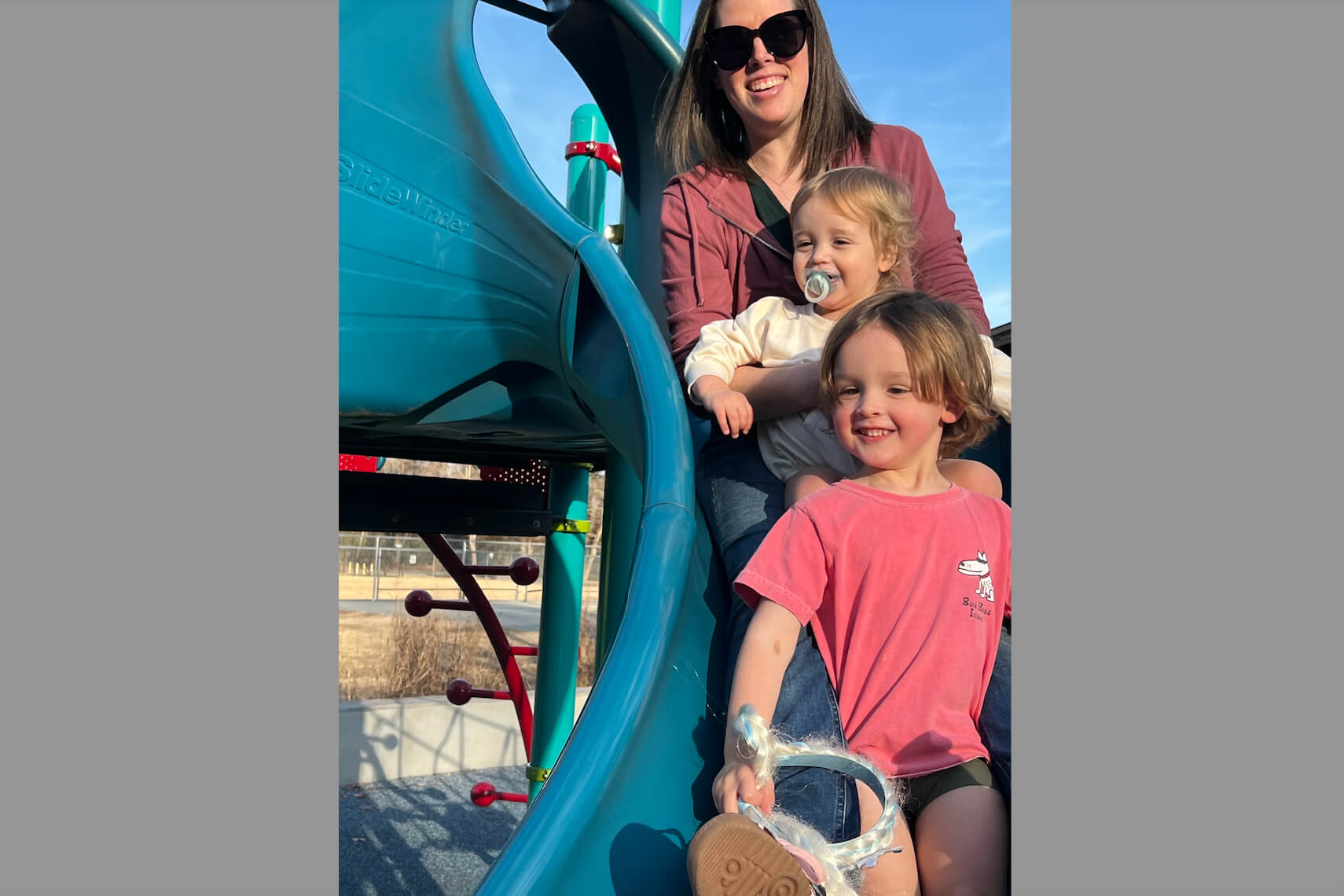 Mother and two young children on a bright blue playground slide outdoors, smiling at the camera (one child has a pacifier).