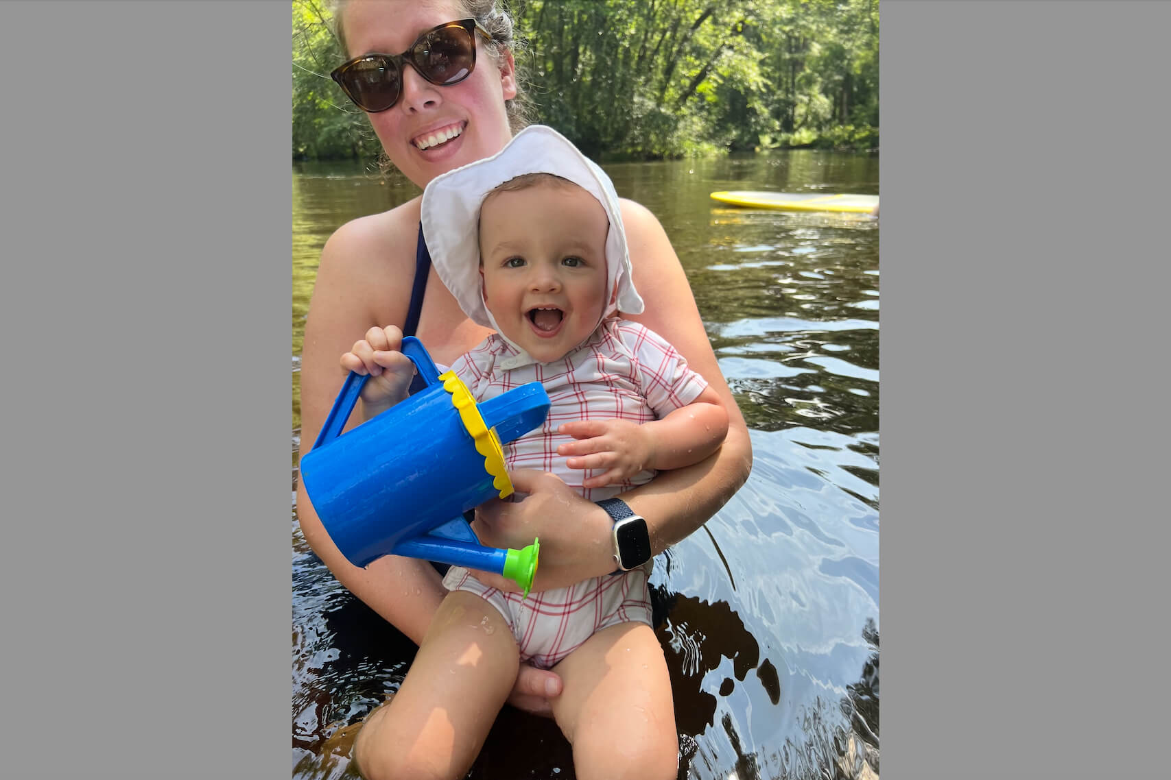 Smiling woman in sunglasses holds a laughing baby wearing a white hat on a lake, the baby gripping a blue toy watering can.