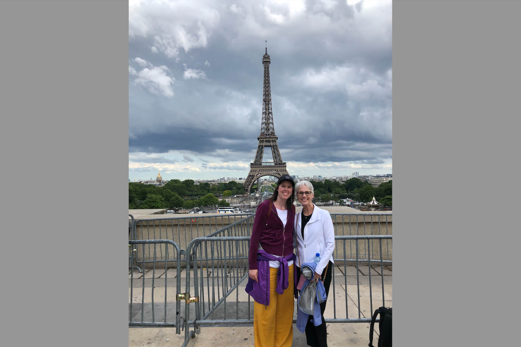 Two women pose for a photo in front of the Eiffel Tower in Paris, with a metal barrier and a cloudy sky behind them, green trees in the background.