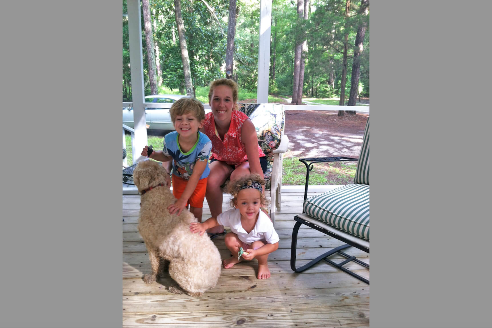 Woman and two children on a wooden porch, with a fluffy beige dog; trees visible in the background.