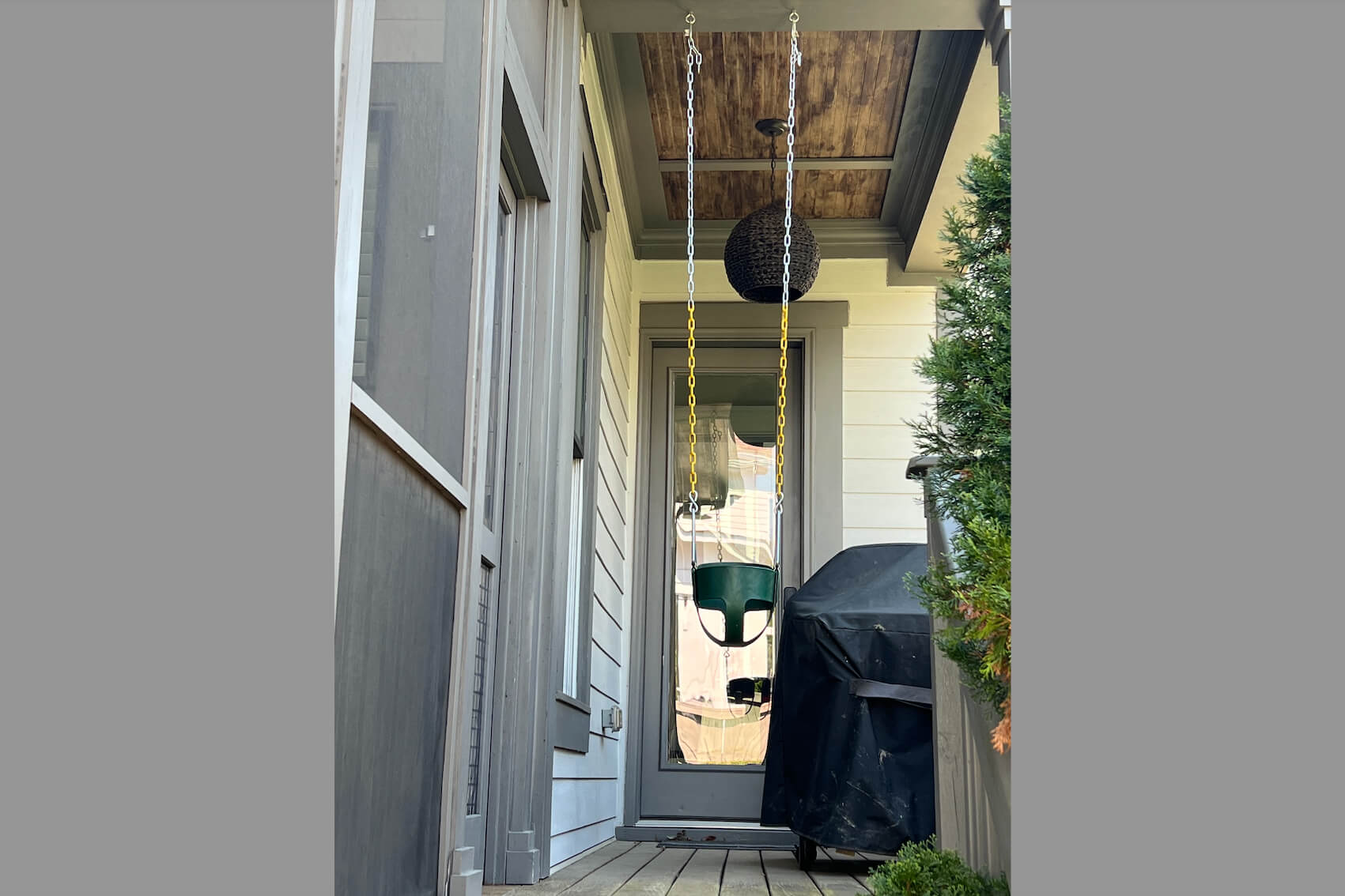 Front porch scene with a green hanging chair suspended by yellow chains, a woven spherical light overhead, and a covered grill to the right.