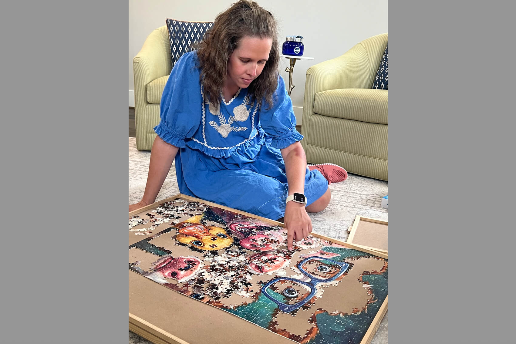 Woman in a blue dress sits on the floor assembling a colorful jigsaw puzzle in a wooden frame.