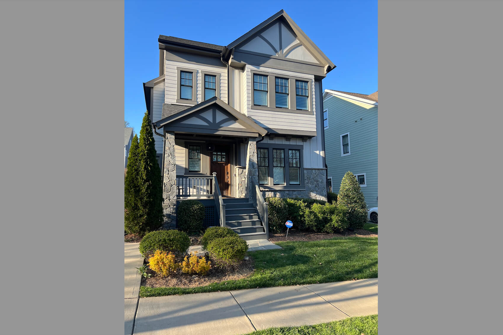 Front view of a two-story craftsman-style house with stone porch columns, gray siding, dark shutters, and a wooden front door amid landscaping.
