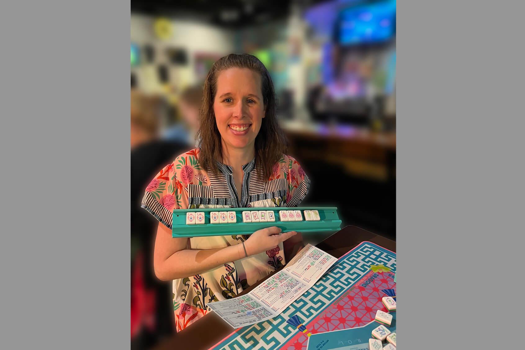 Smiling woman in a floral top holds a turquoise Mahjong tile rack, with a colorful game mat and papers on the table in front.