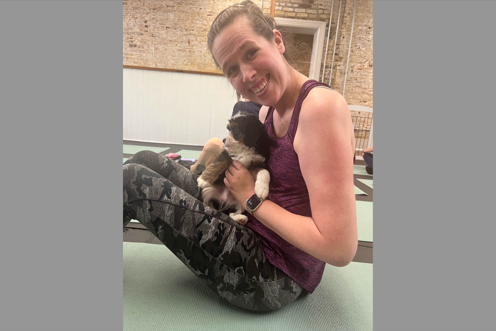 Woman in a purple tank top sits on a yoga mat, smiling while holding a small fluffy puppy during a workout session in a gym-like space.
