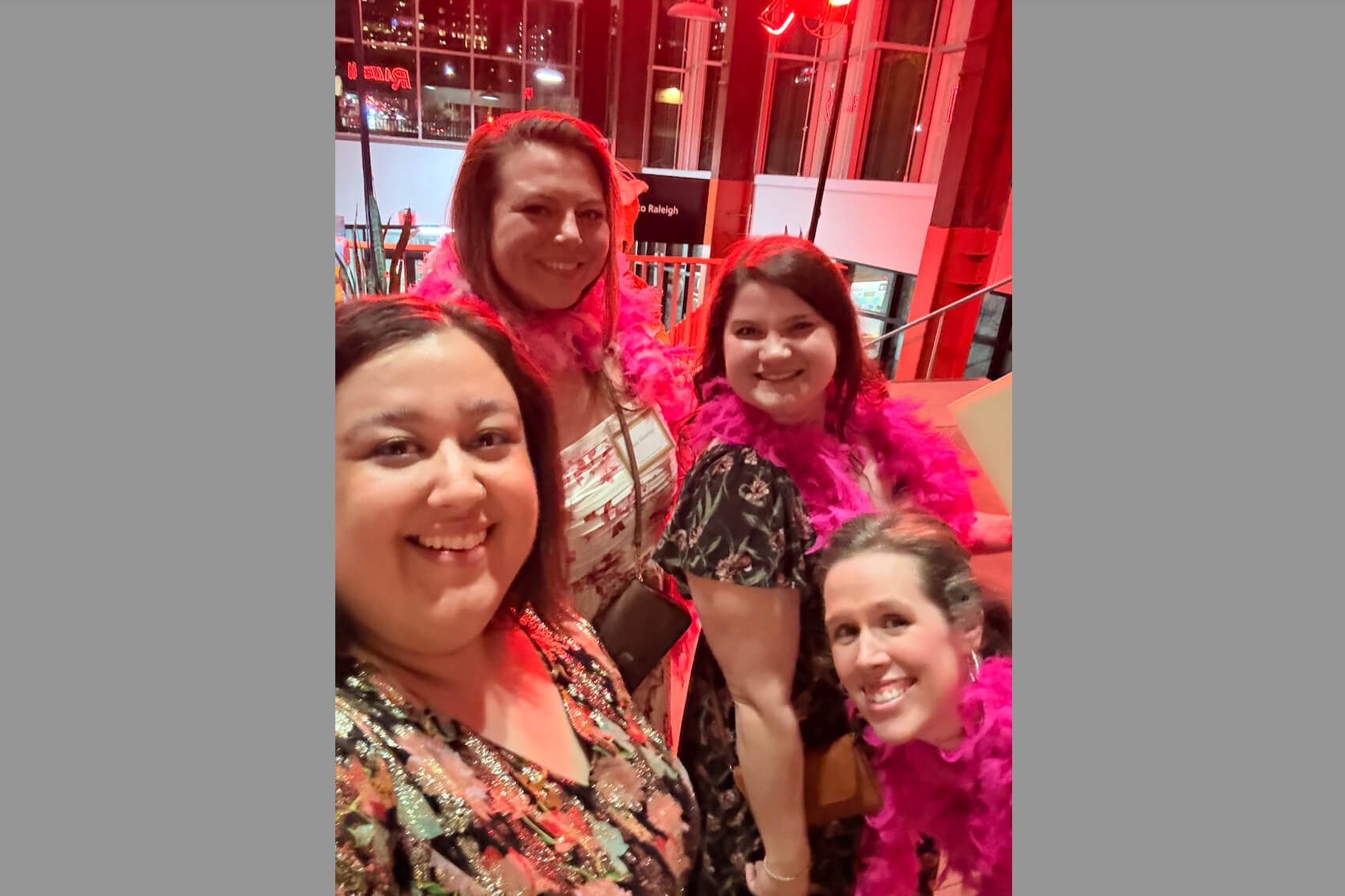Four women pose for a group photo at a festive event, wearing colorful outfits and pink feather boas under red lighting.