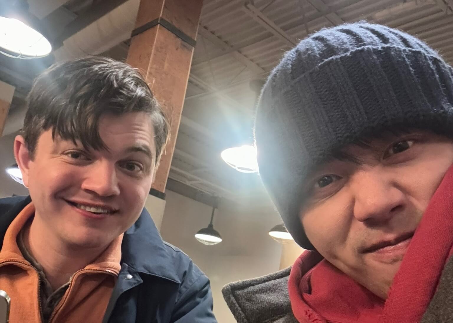 Two men taking a casual indoor selfie; one in a navy jacket with an orange fleece, the other wearing a gray knit beanie and red scarf, both smiling.—indoor setting with wooden columns and hanging lights