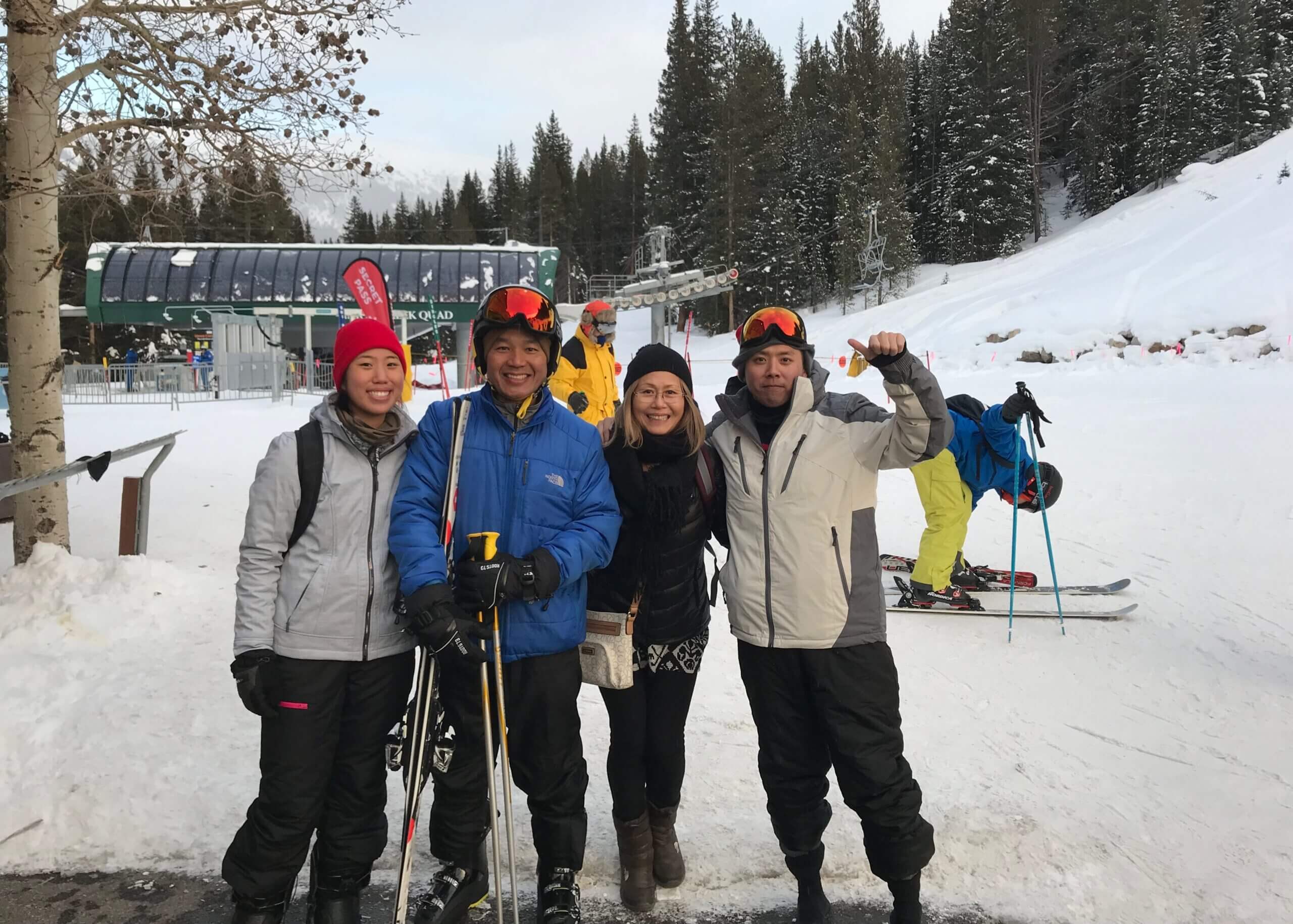 Four friends in ski gear posing for a group photo on a snowy slope with trees and a ski lift in the background.