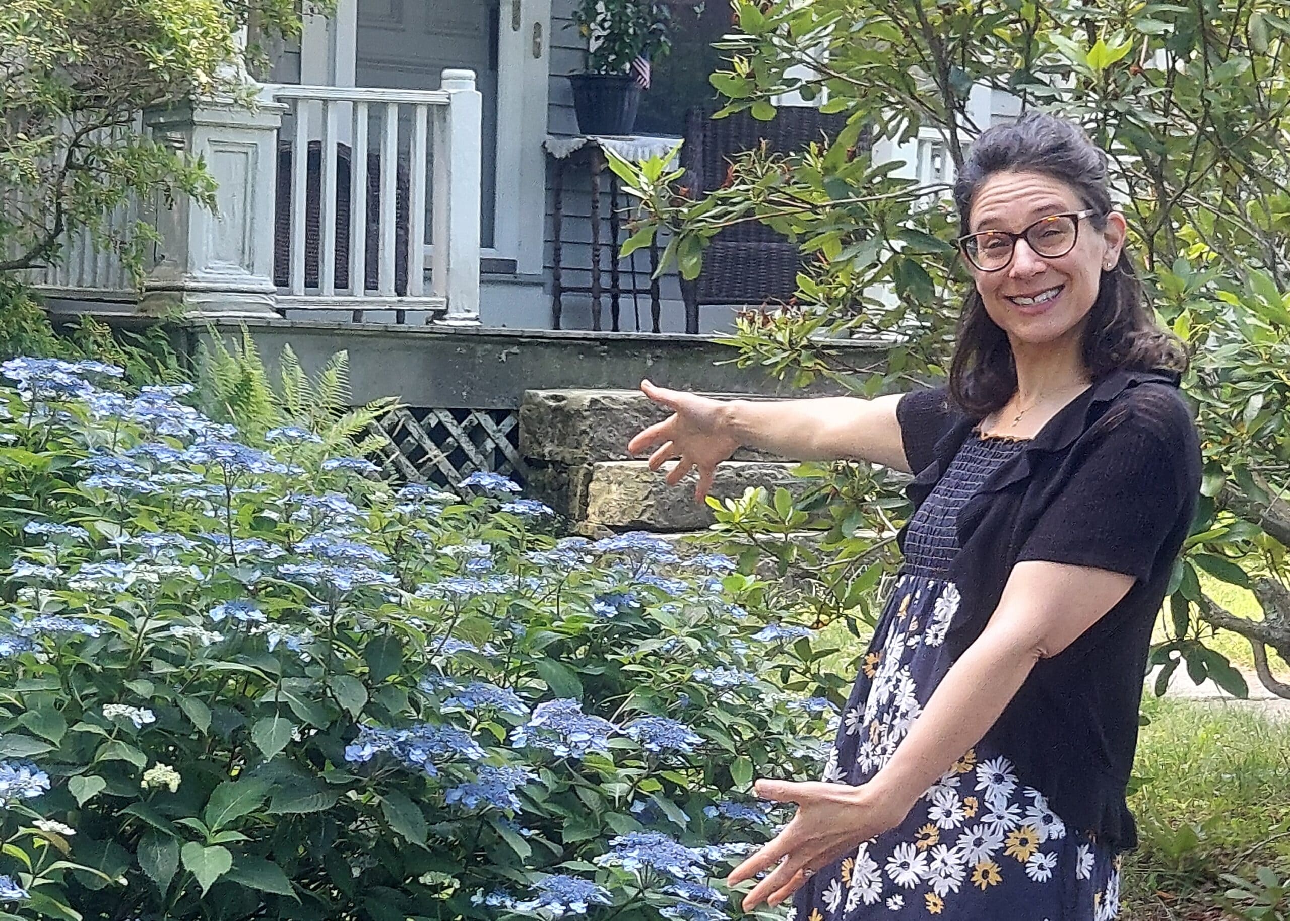 Smiling woman with glasses stands in a garden among blue hydrangeas, arms extended as if pointing or gesturing to the left, with a porch in the background.