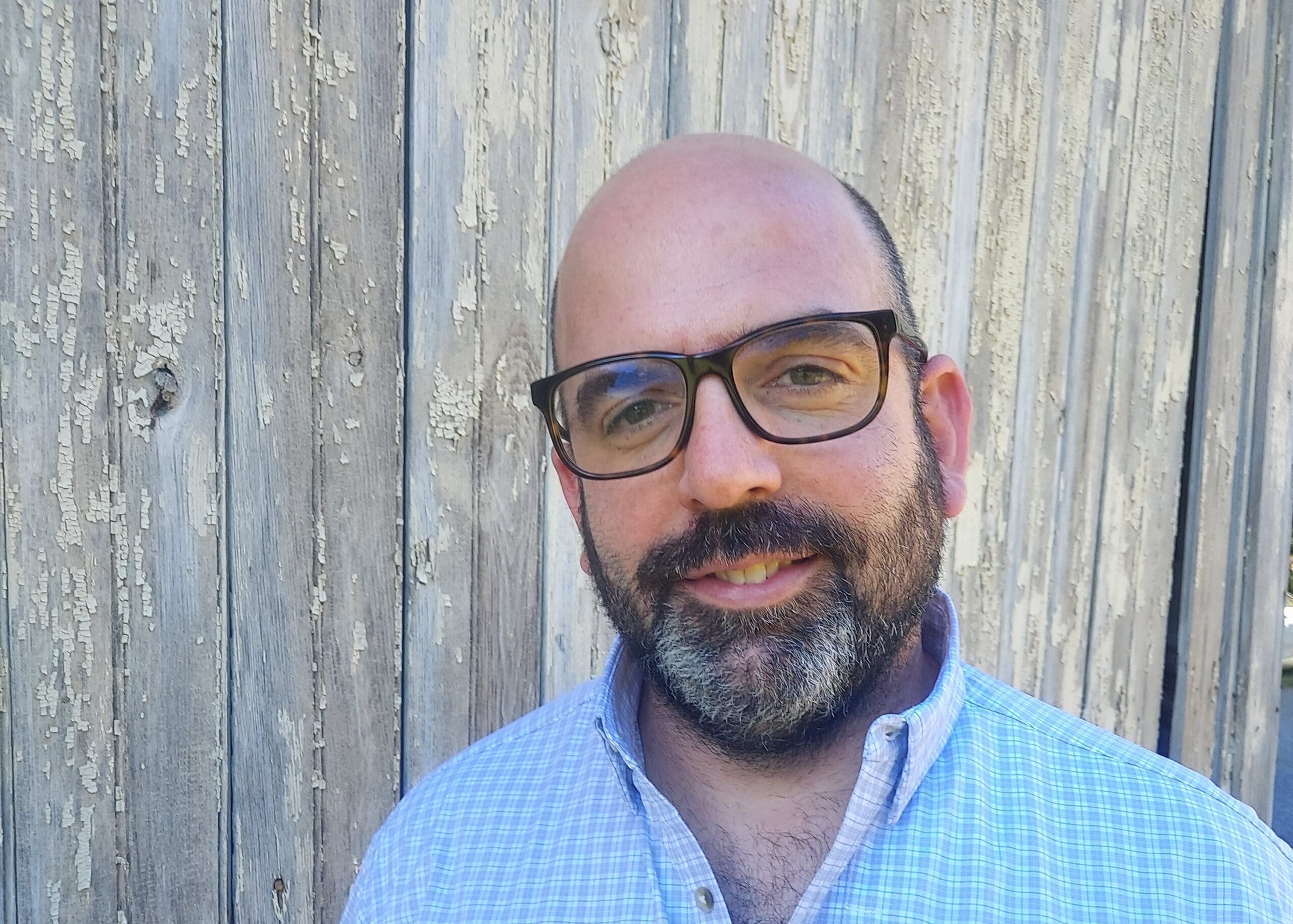 Self-portrait of a man with glasses and a beard smiling against a weathered wooden fence. Head and shoulders visible in daylight.