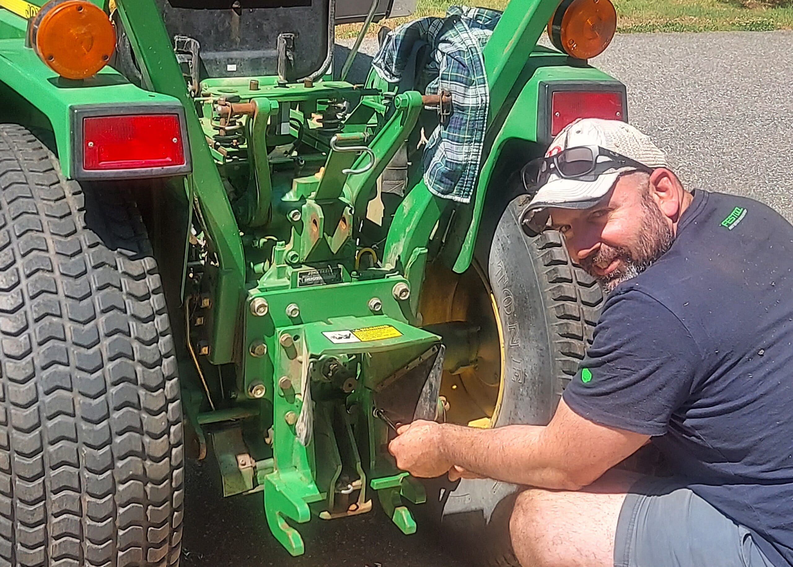 Man wearing a cap and sunglasses crouches beside a green tractor, inspecting the hitch near a large rear tire.