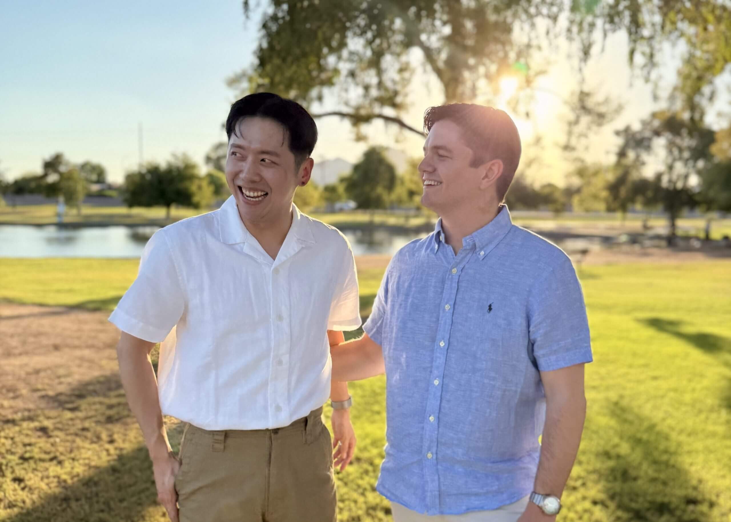 Two men standing in a sunlit park, smiling at each other; one wears a white shirt, the other a light blue shirt near a pond.