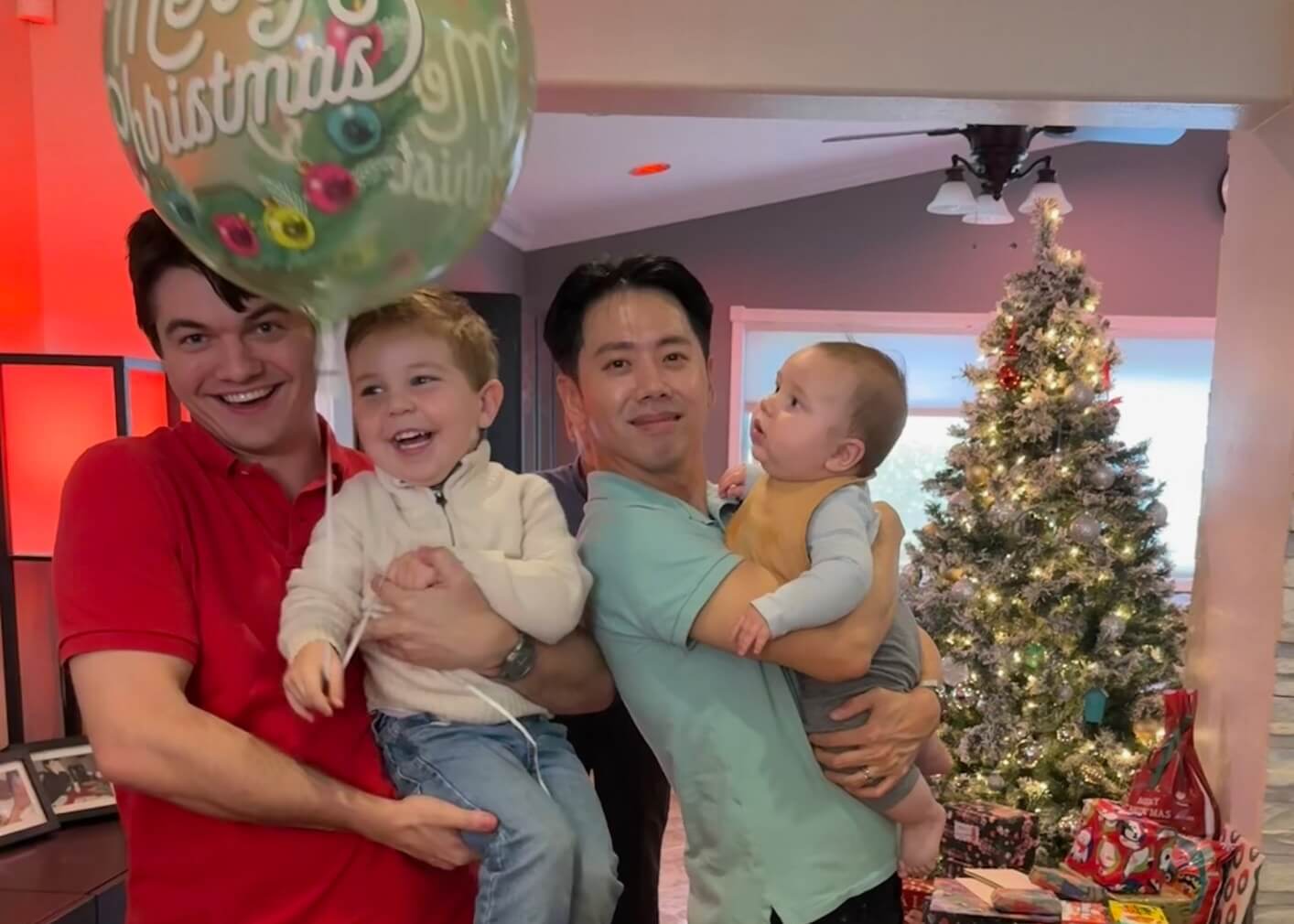 Two men holding smiling children in a cozy living room with a Christmas tree and wrapped gifts in the background.