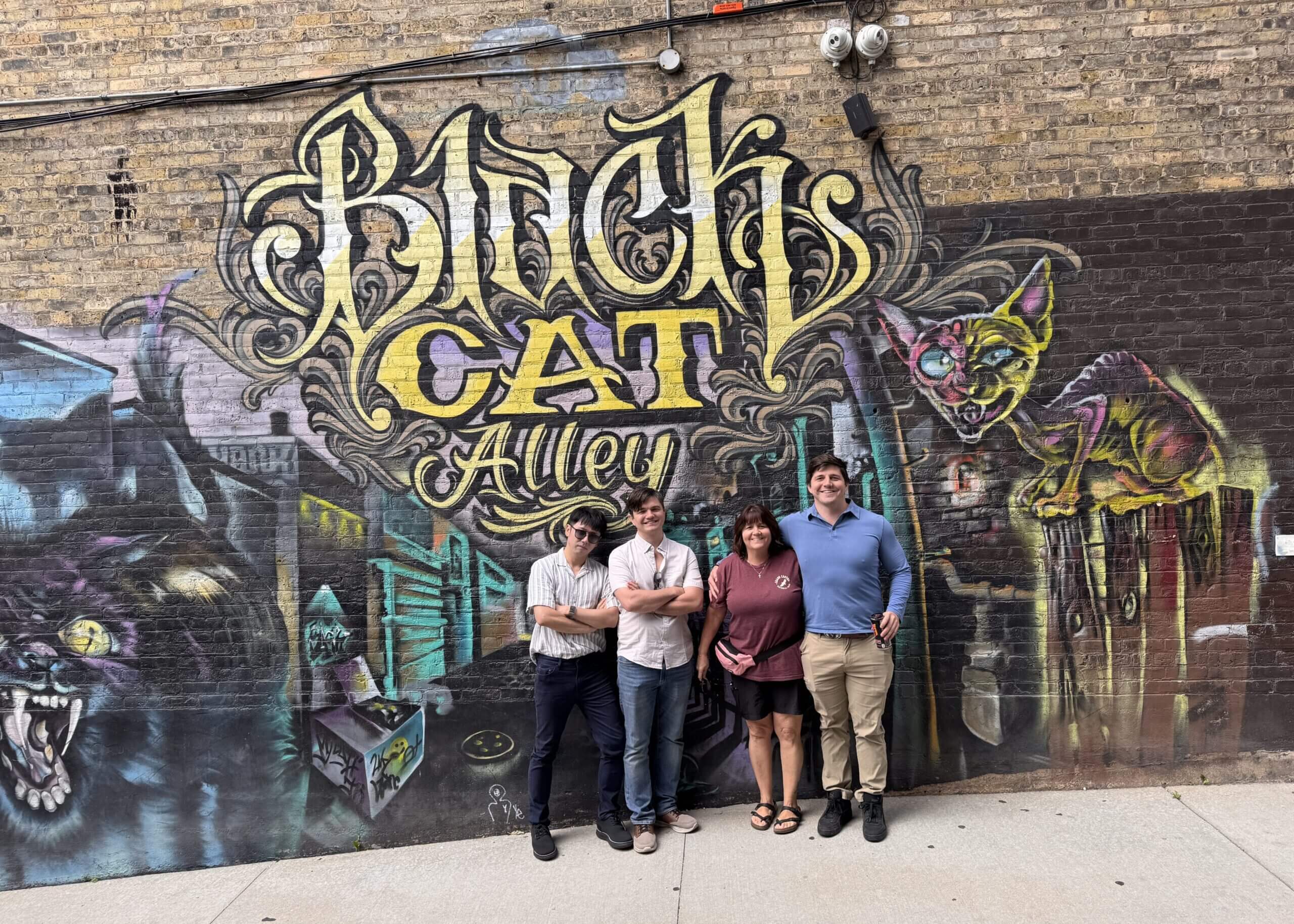 Five people pose together in front of a vivid graffiti mural on a brick wall, smiling at the camera.
