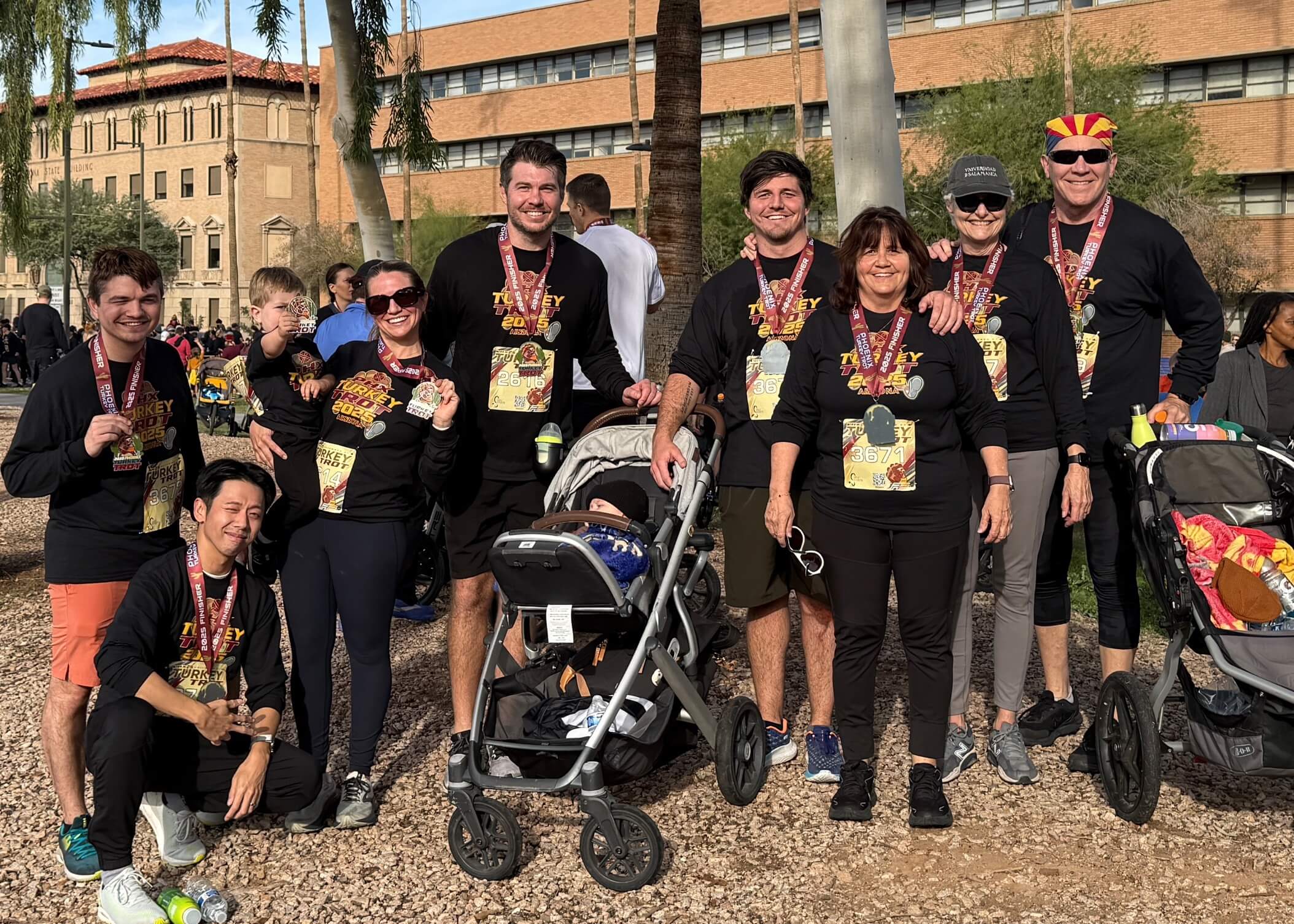 Group of marathon runners posing for a photo outdoors, wearing matching black shirts and race bibs with medals around their necks, some standing and one in a stroller-style wheelchair.