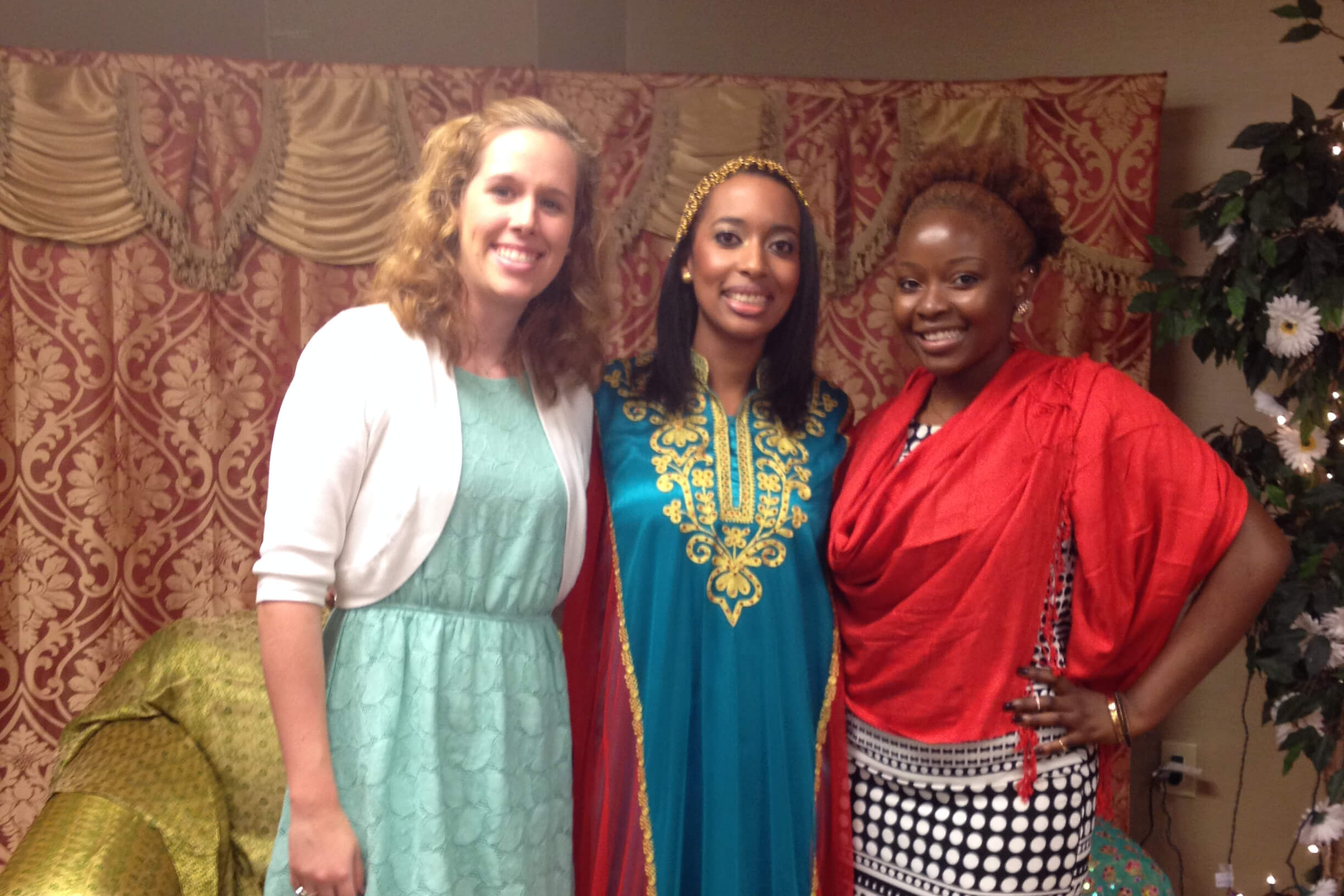 Three women pose together indoors, smiling, wearing colorful dresses against a patterned backdrop.