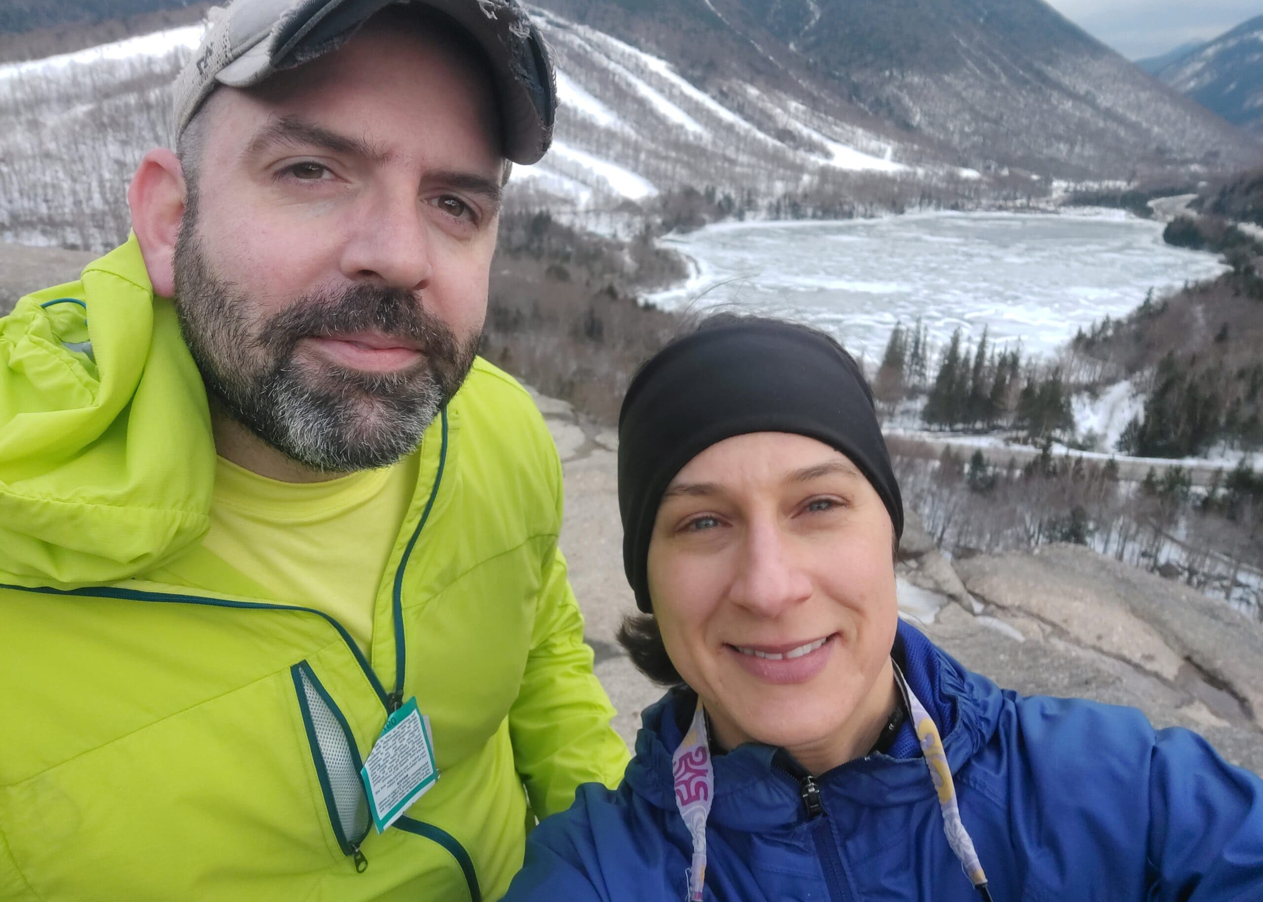 Selfie of a man in a bright lime jacket and a woman in a blue jacket, with snow-covered mountains and a frozen river behind them.