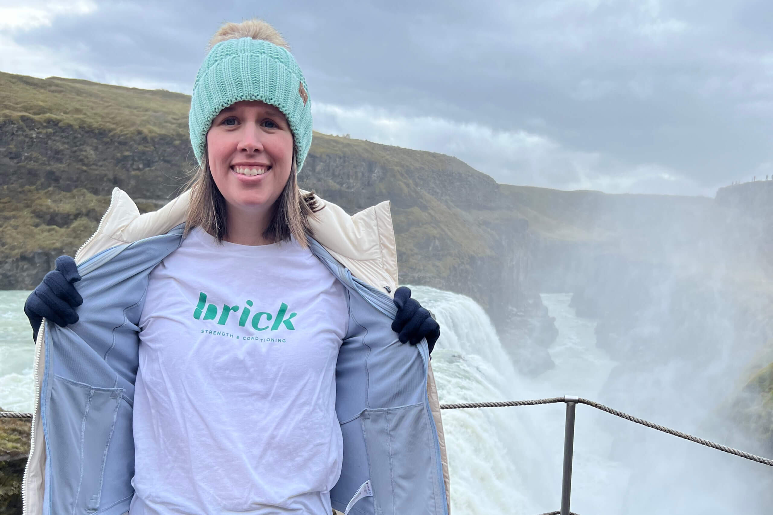 Smiling person wearing a teal knit hat with a pom and light jacket, in front of a misty waterfall and rocky cliffs.
