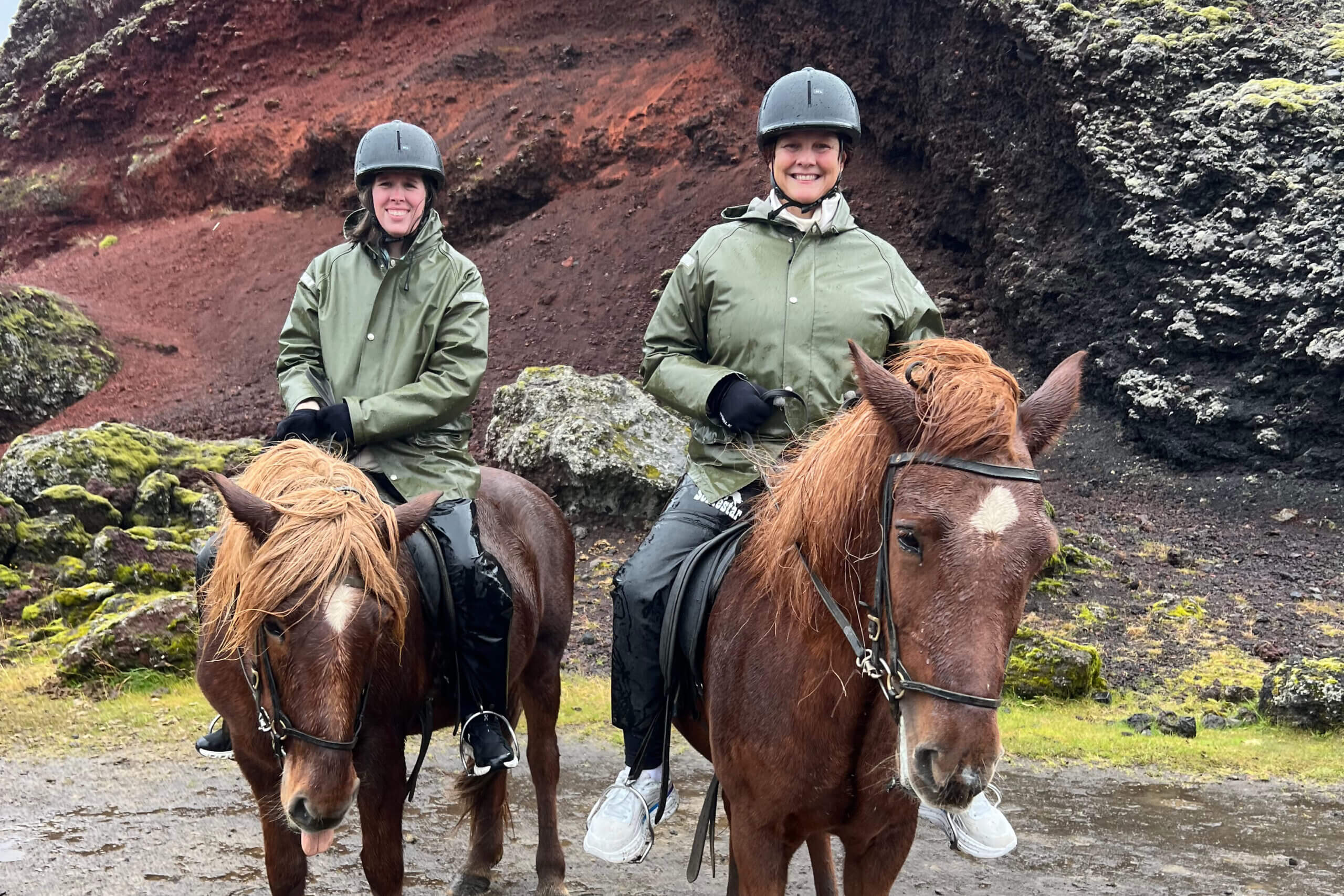Two riders on brown horses wearing helmets and green jackets, posing on a rocky, mossy trail outdoors behind them the red-brown volcanic rock wall.