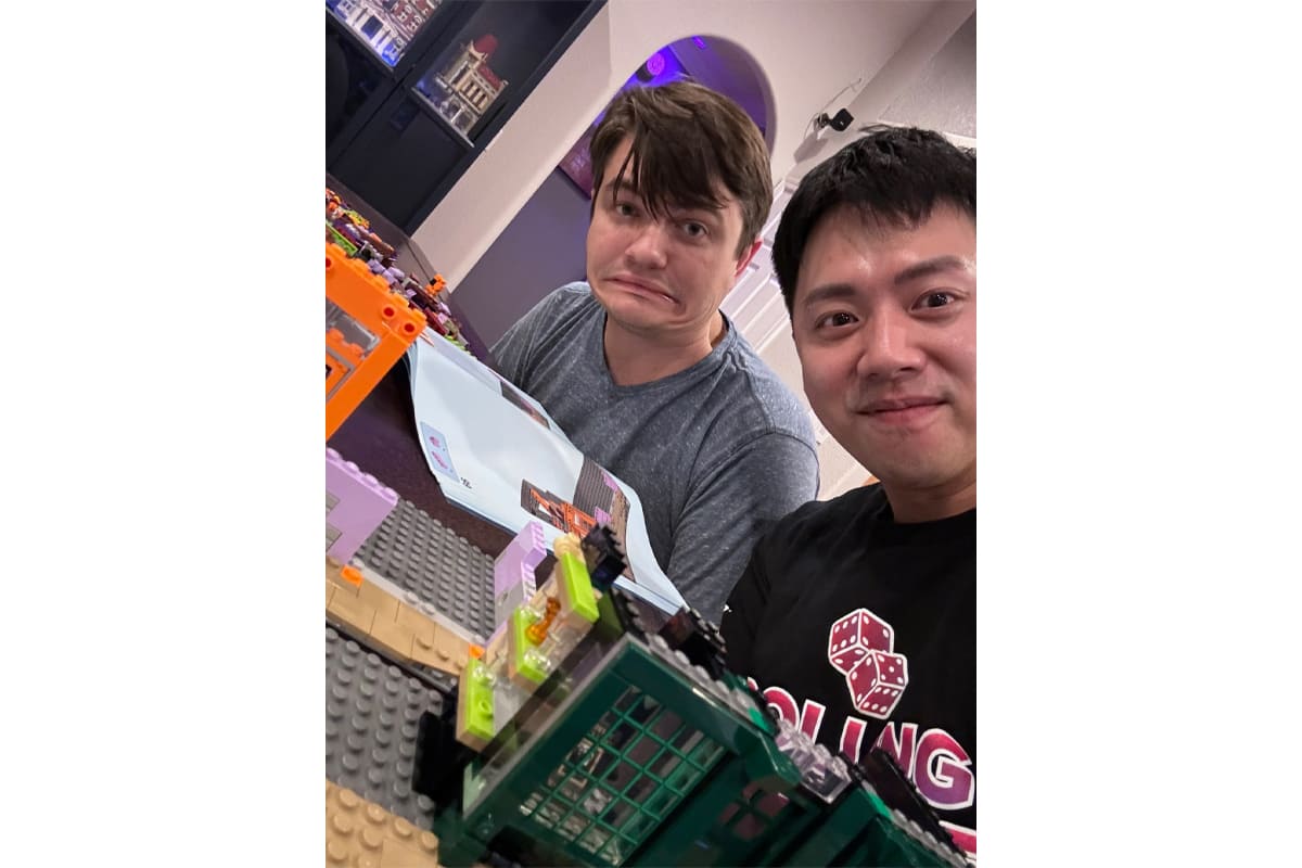Two men pose with a large, colorful LEGO build, one smiling and the other making a funny grimace at the camera.