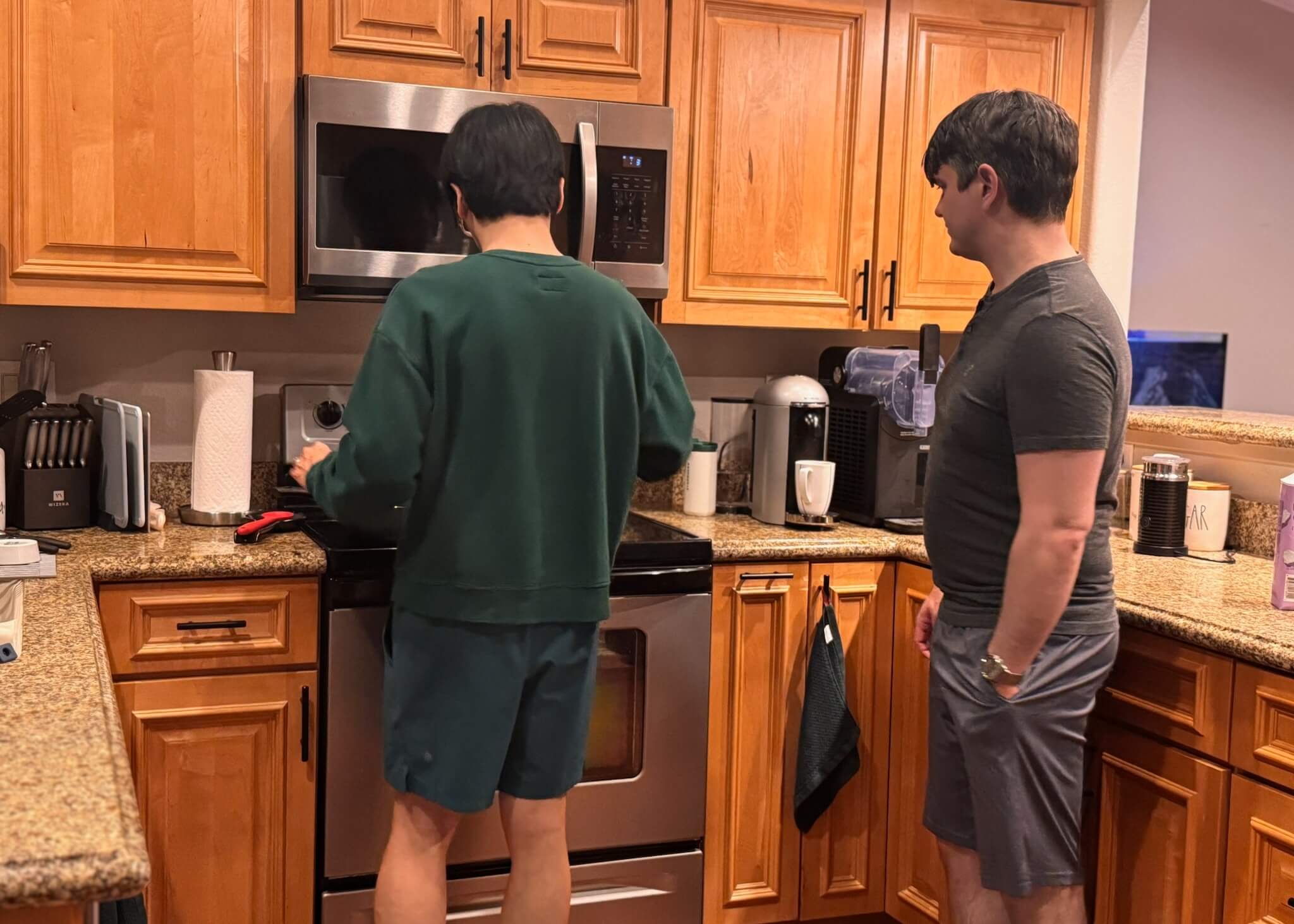 Two people in a kitchen: one in green scrubs stands at a stainless steel oven while the other observes nearby.